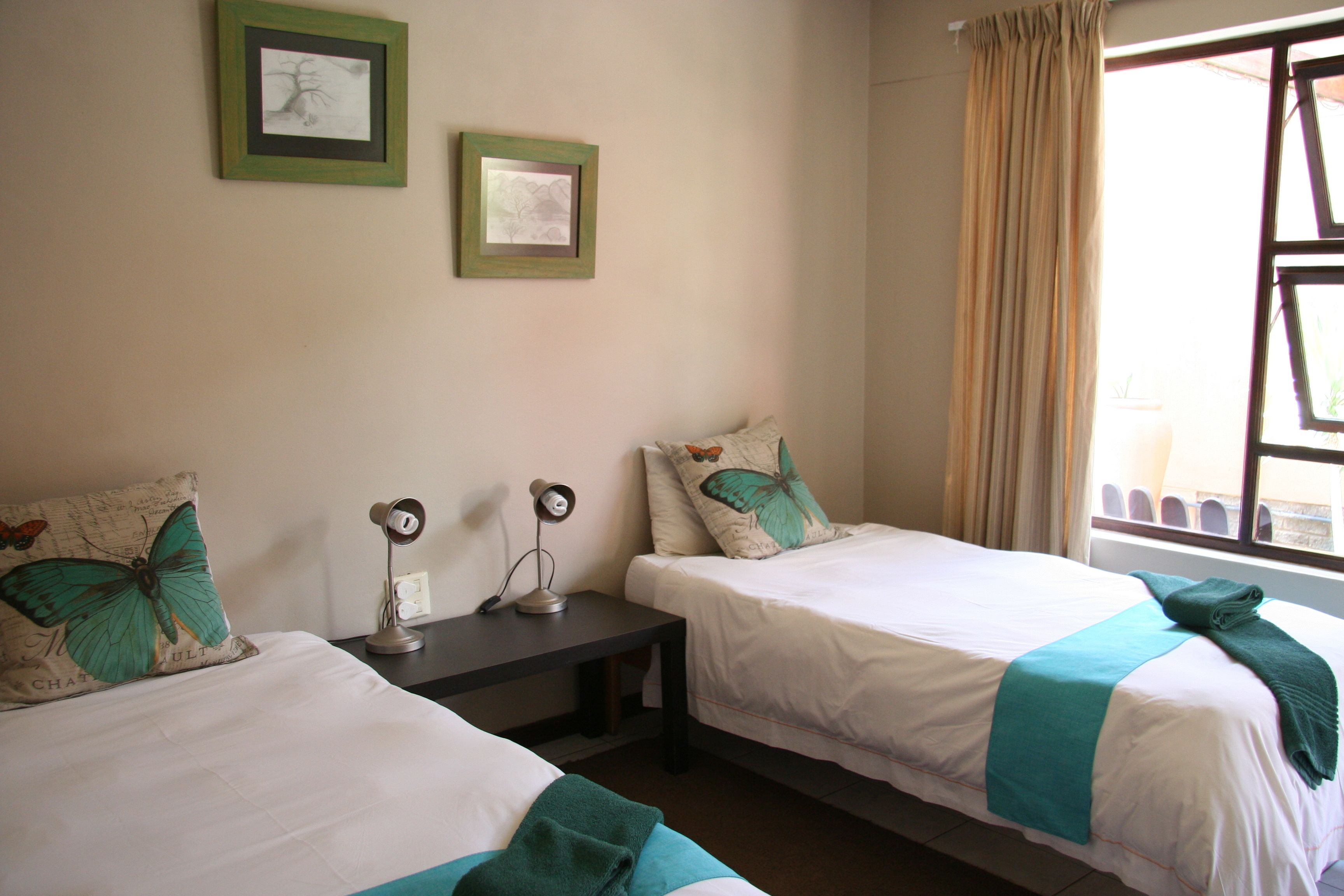 Two single beds with matching green and white bedding, a small bedside table with a lamp, and two framed pictures on the wall.