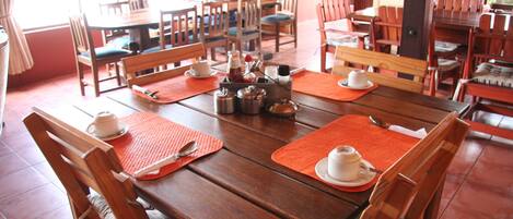 A dining area with wooden tables and chairs, orange placemats, and white cups and saucers.