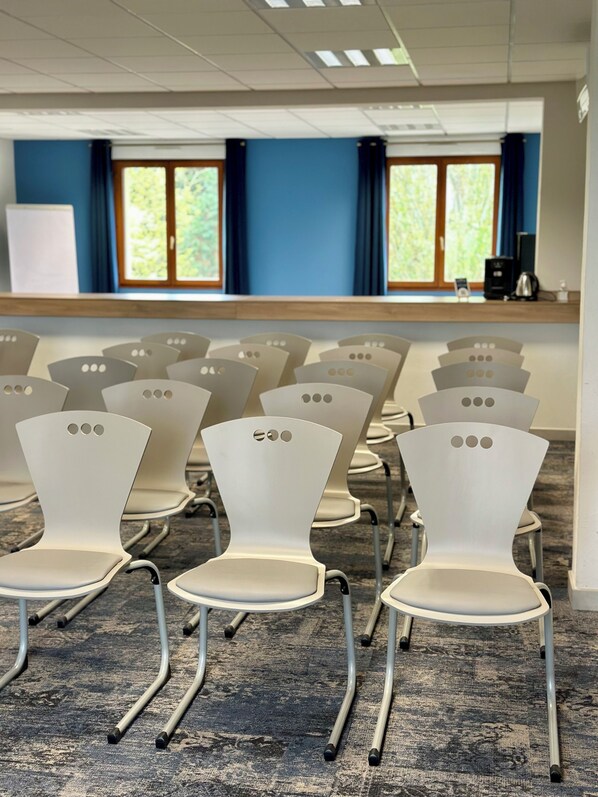 Un salón de conferencias con filas de sillas blancas, piso alfombrado y un ventanal con cortinas azules.