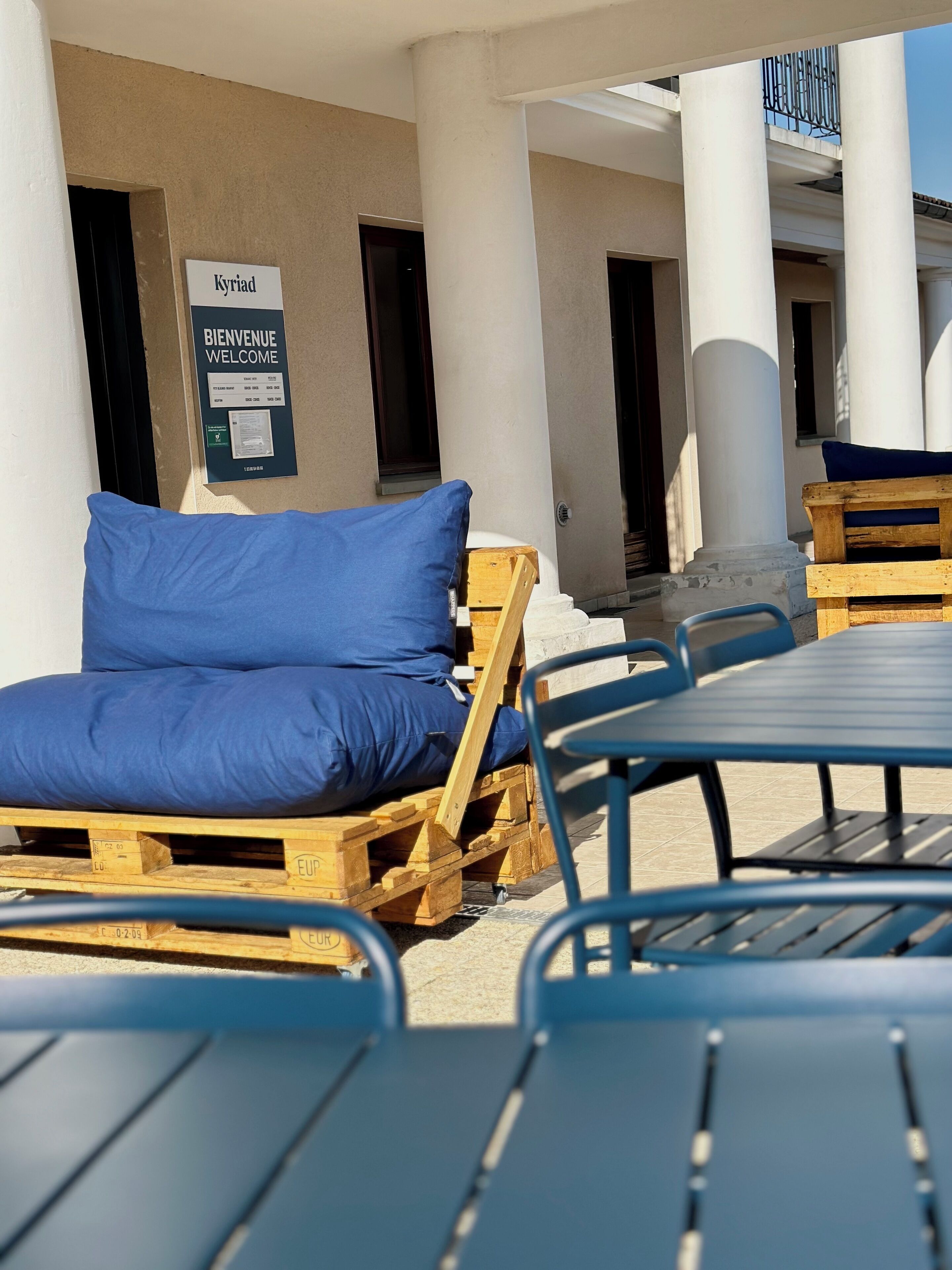 Entrada de hotel con un cartel que dice 'Welcome', asientos exteriores con cojines azules y muebles de madera.