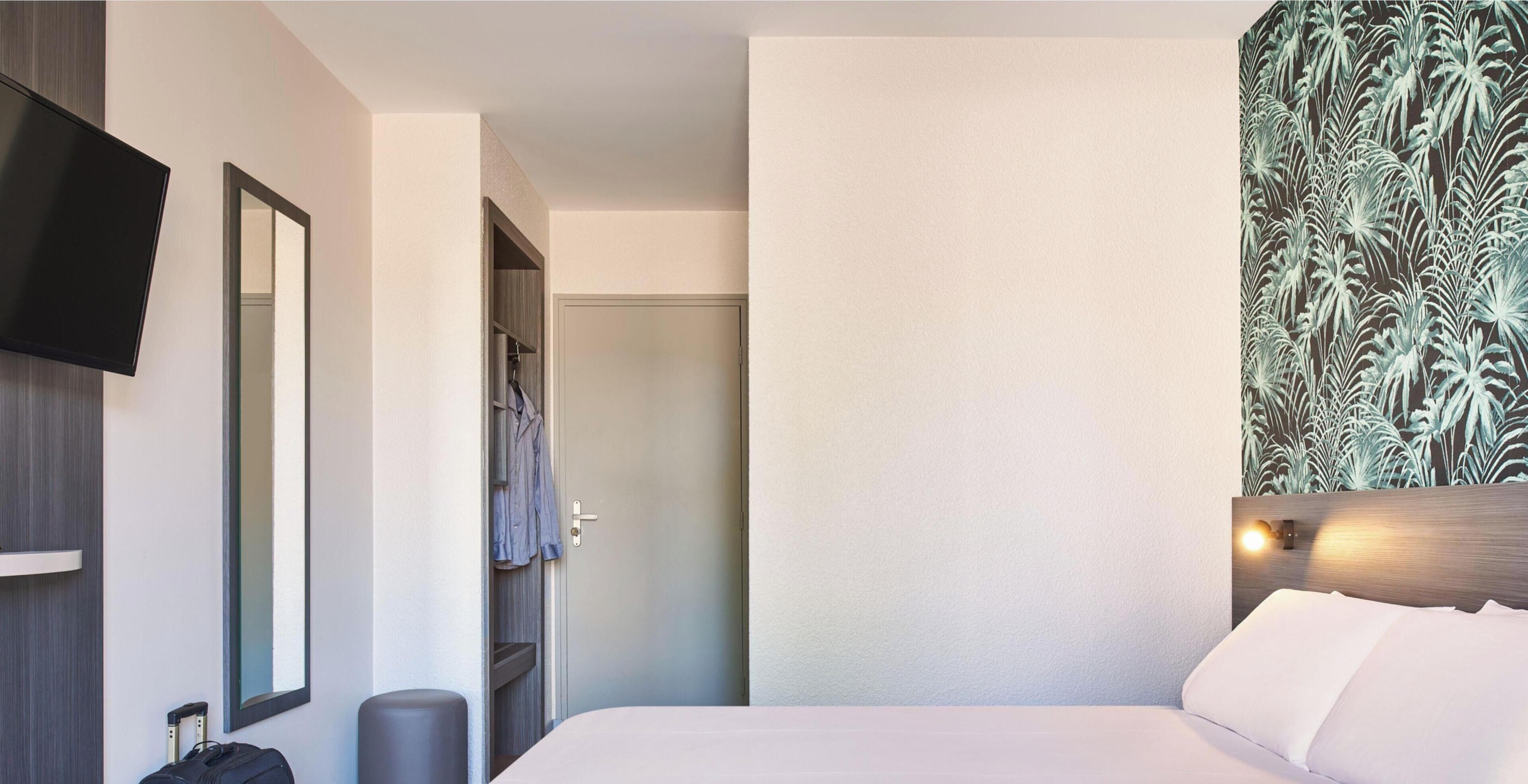 A hotel room with a bed, a television mounted on the wall, a wardrobe, and a door leading to a bathroom.
