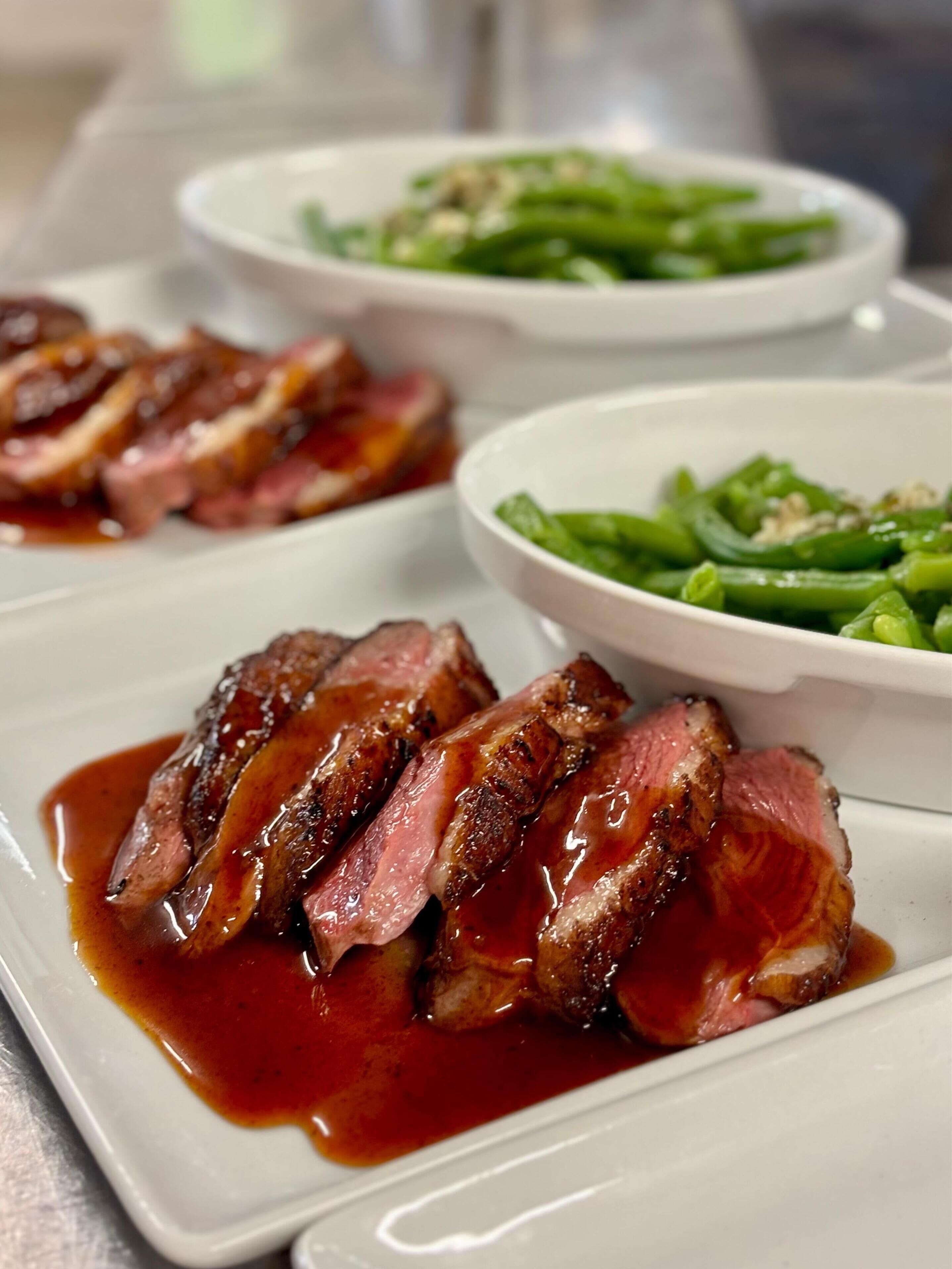 Une assiette de tranches de steak nappées d’une sauce onctueuse, accompagnée d’un bol de haricots verts et d’un autre plat visible à l’arrière-plan.