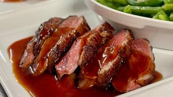 Une assiette de tranches de steak nappées d’une sauce onctueuse, accompagnée d’un bol de haricots verts et d’un autre plat visible à l’arrière-plan.