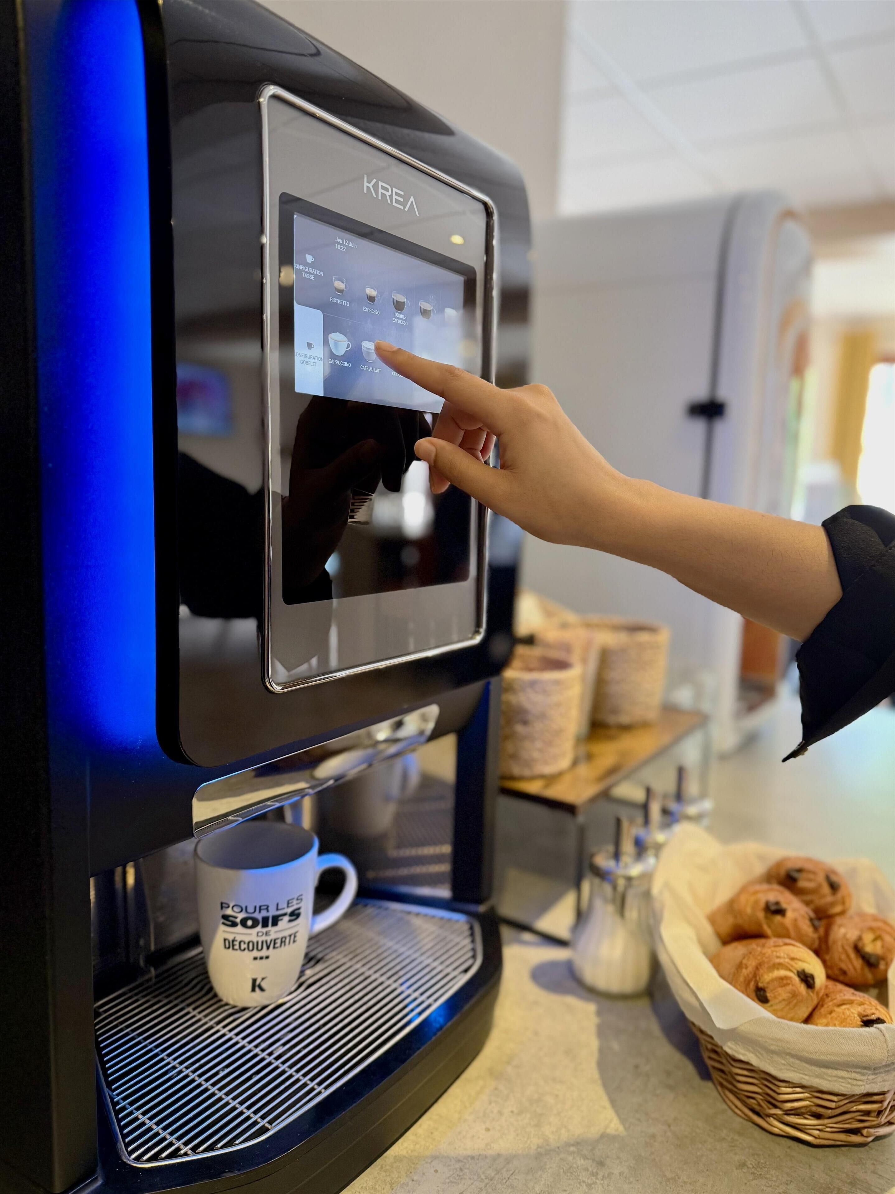 Una cafetera KREA con pantalla táctil, una taza de café y una cesta de croissants sobre la encimera.