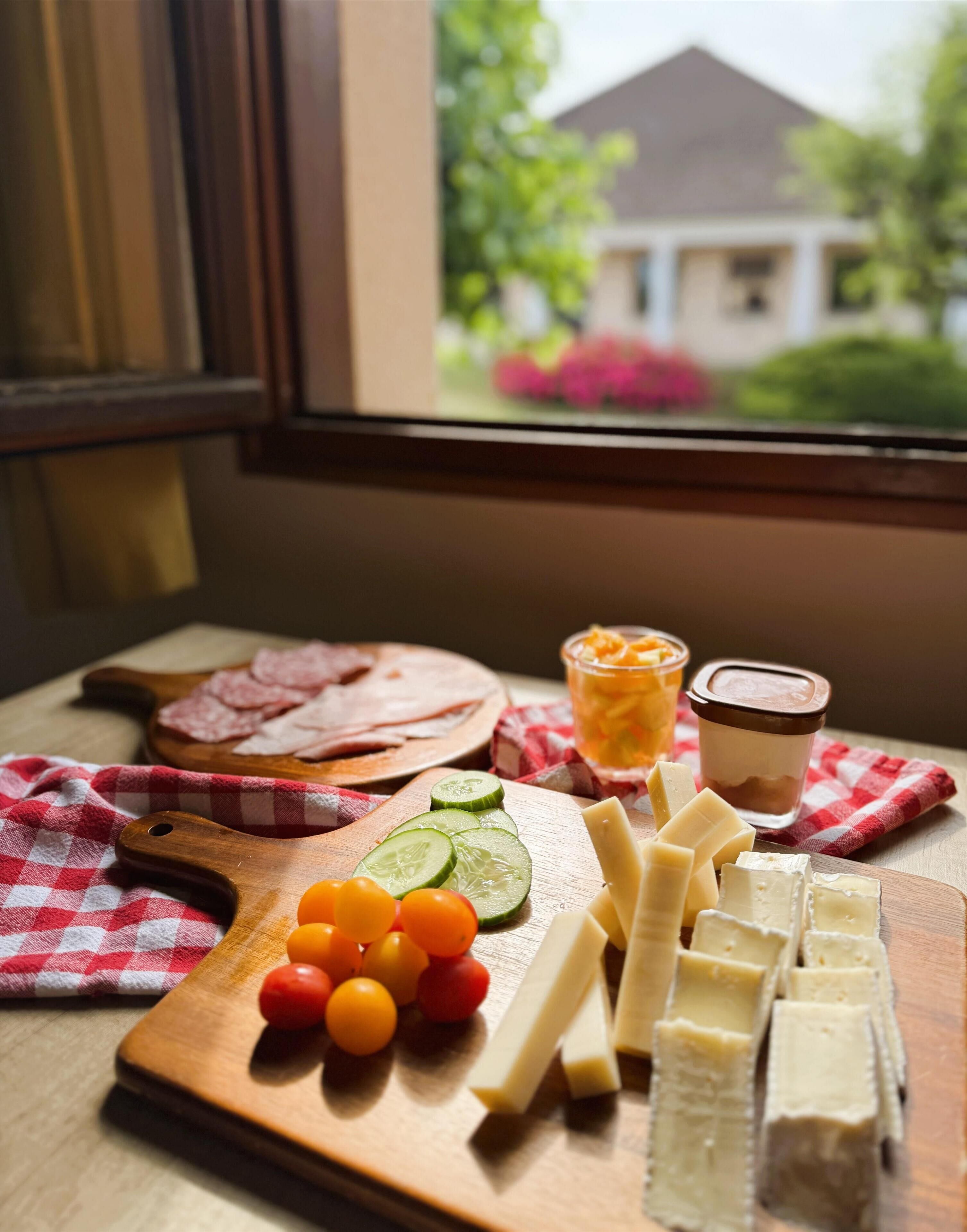 Une planche en bois sur laquelle sont disposés du fromage, des tomates et des concombres, posée sur une table près d’une fenêtre donnant sur une maison et un jardin.