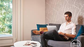 Un homme est assis sur un canapé gris, avec un ordinateur portable, une tasse de café et un smartphone posés sur une petite table ronde à côté de lui.