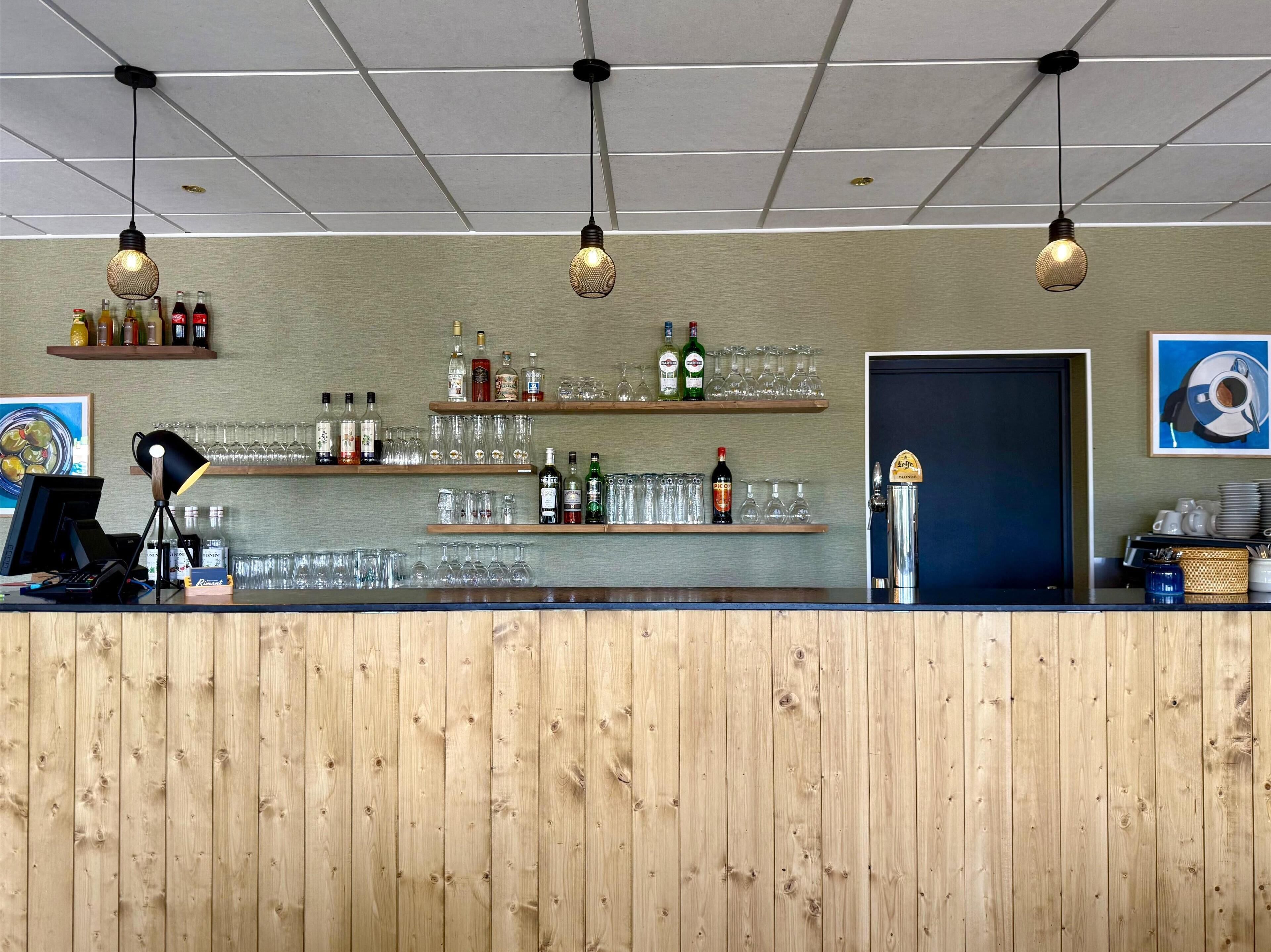 Zona de bar con paneles de madera, estantes con botellas y vasos, y lámparas colgantes.