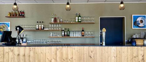Zona de bar con paneles de madera, estantes con botellas y vasos, y lámparas colgantes.
