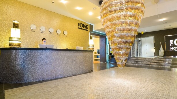 Un espace d’accueil d’hôtel au design moderne, comprenant un comptoir d’accueil, un grand luminaire décoratif suspendu et un mur orné d’horloges.