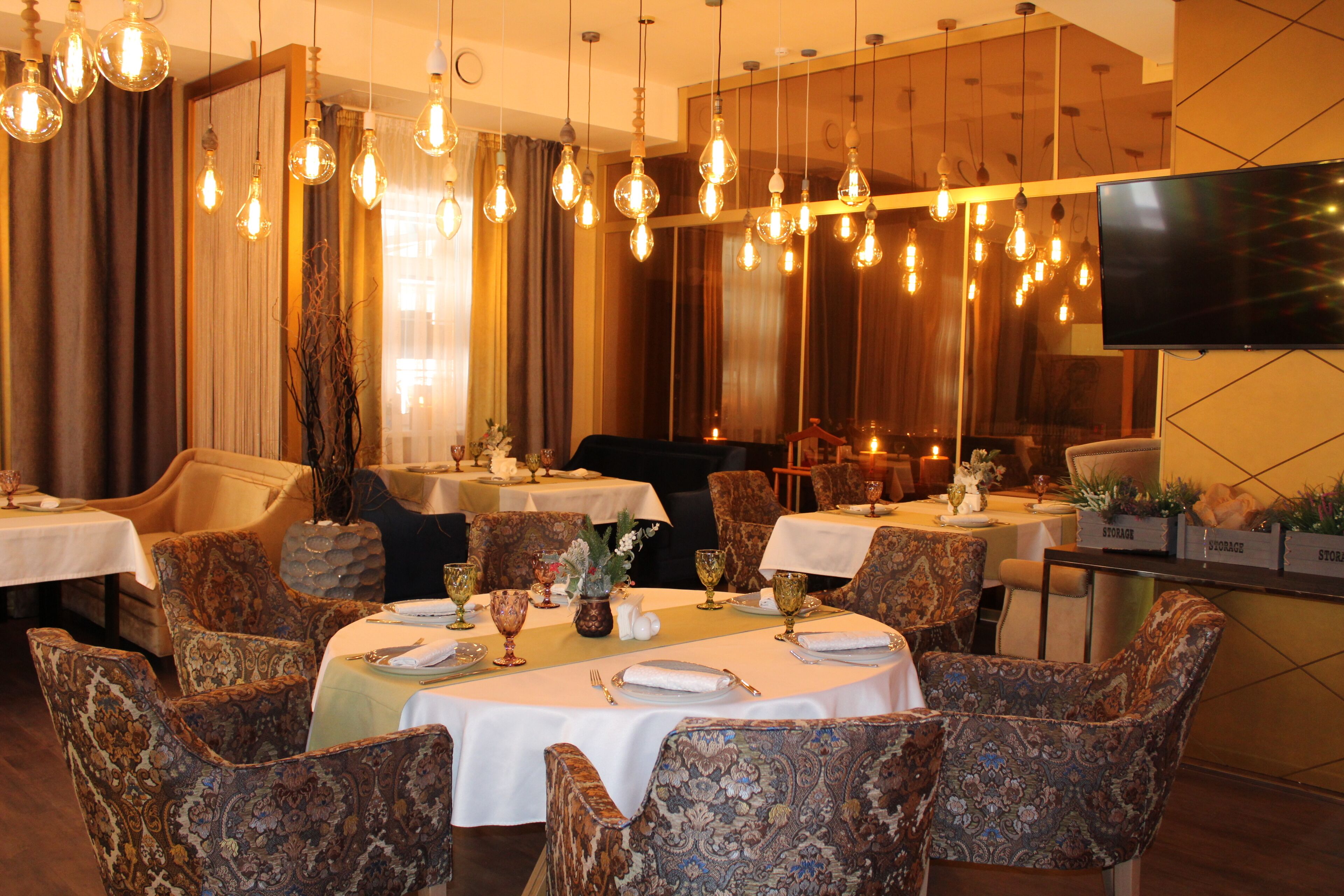 A restaurant with tables set for dining, patterned chairs, hanging light bulbs, and a television on the wall.