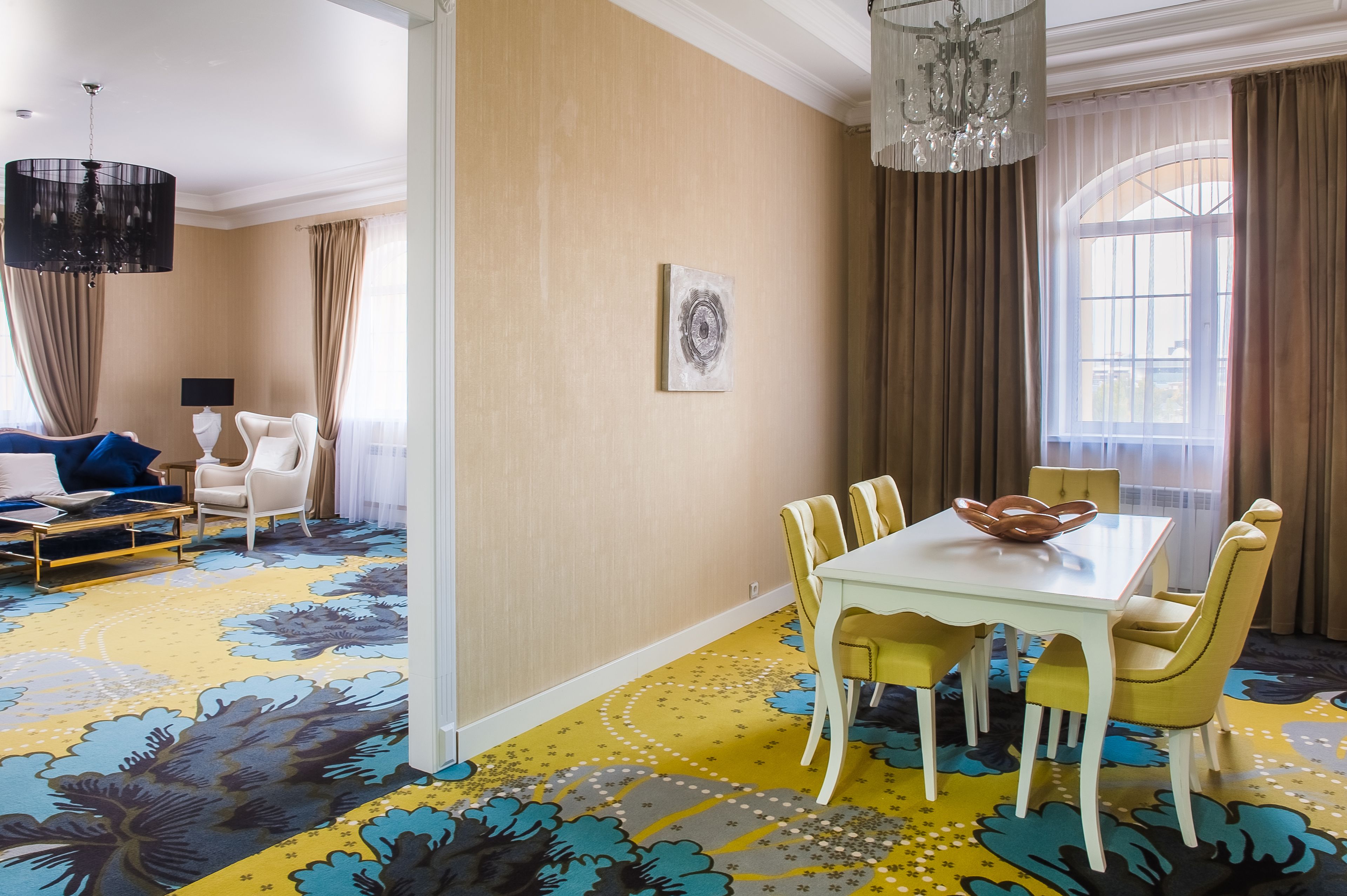 A hotel room with a dining area featuring a white table and yellow chairs, a large floral carpet, and a chandelier.