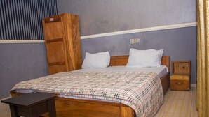 A bedroom with a wooden bed, two pillows, a nightstand, a wooden wardrobe, and a small table. - Duckland Hotel and Suites Ltd (Ikorodu)