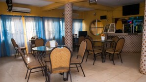 A dining area with multiple tables and chairs, a bar counter, and a television mounted on the wall. - D Jos Hotel (Ijede)