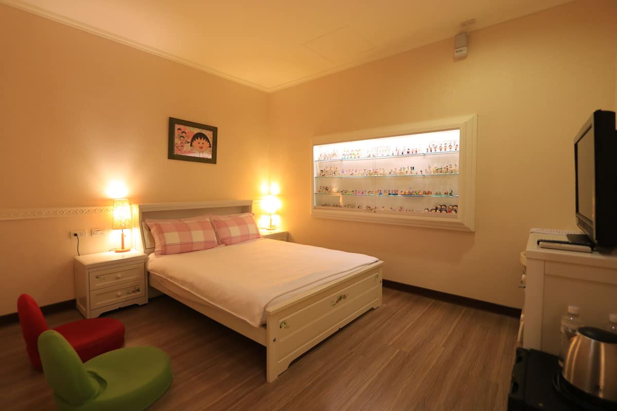 Une chambre à coucher comprenant un lit, des tables de chevet, une commode, une fenêtre avec des étagères, un téléviseur et un fauteuil rouge.