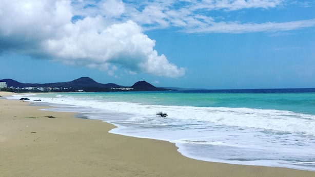 Una playa con aguas turquesas cristalinas, una orilla arenosa y montañas distantes bajo un cielo parcialmente nublado.