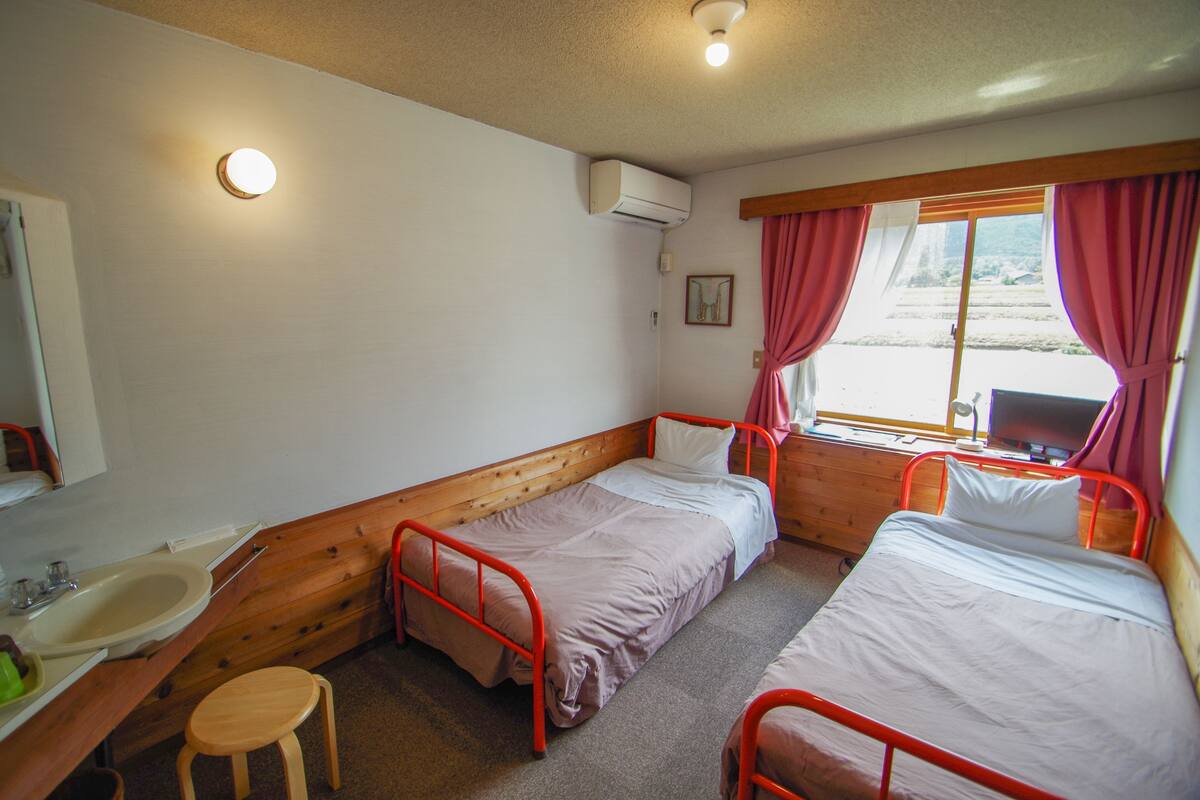 A room with two single beds, a wooden bench, a sink, and a window with curtains.