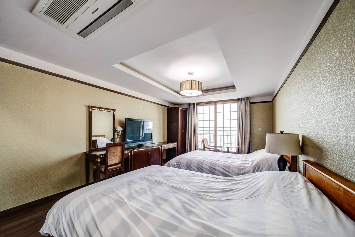 A hotel room with two beds, a desk with a television, a lamp, and a large window with curtains.