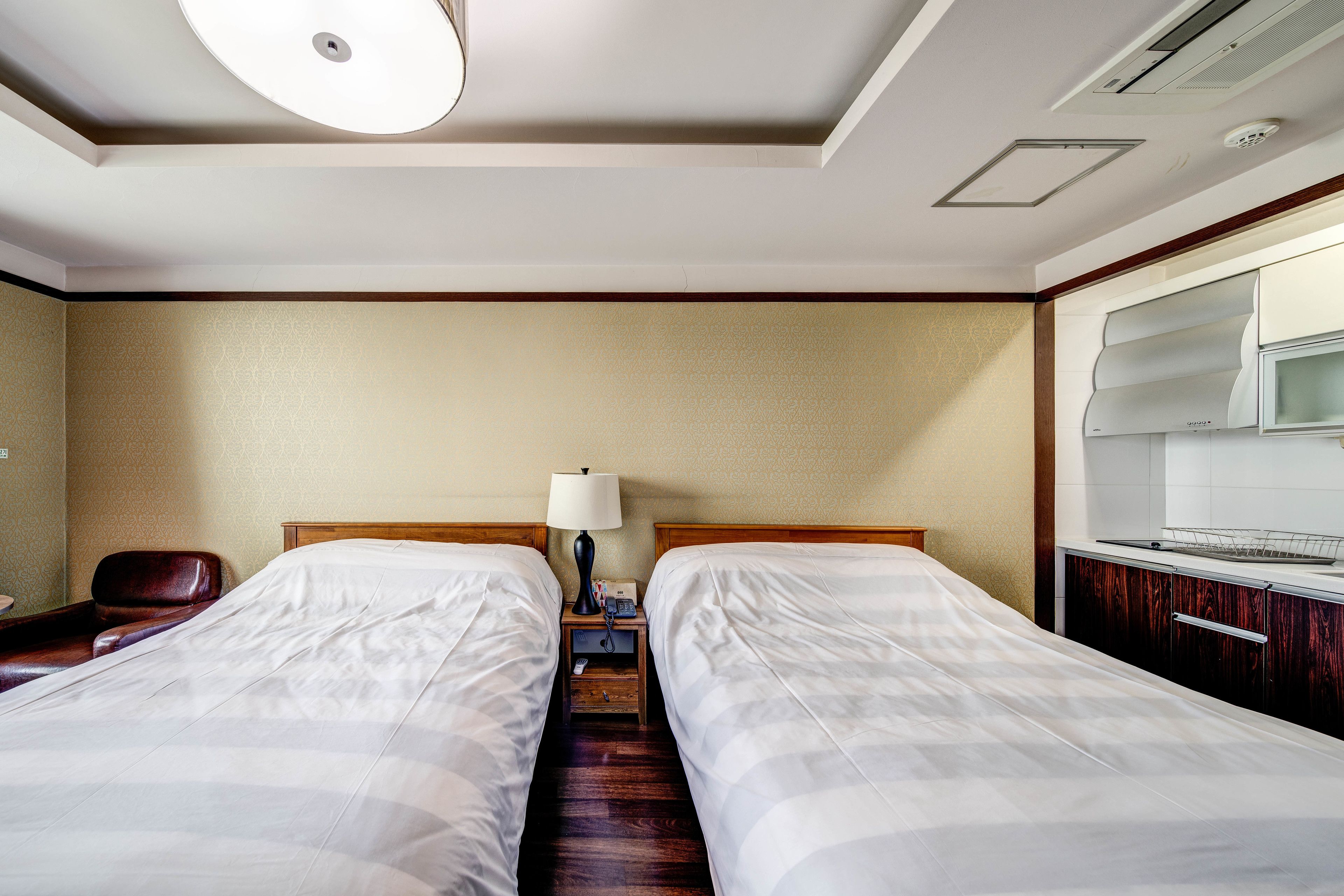 A hotel room with two beds, a desk with a chair, a lamp, and a kitchenette.