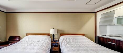 A hotel room with two beds, a desk with a chair, a lamp, and a kitchenette.