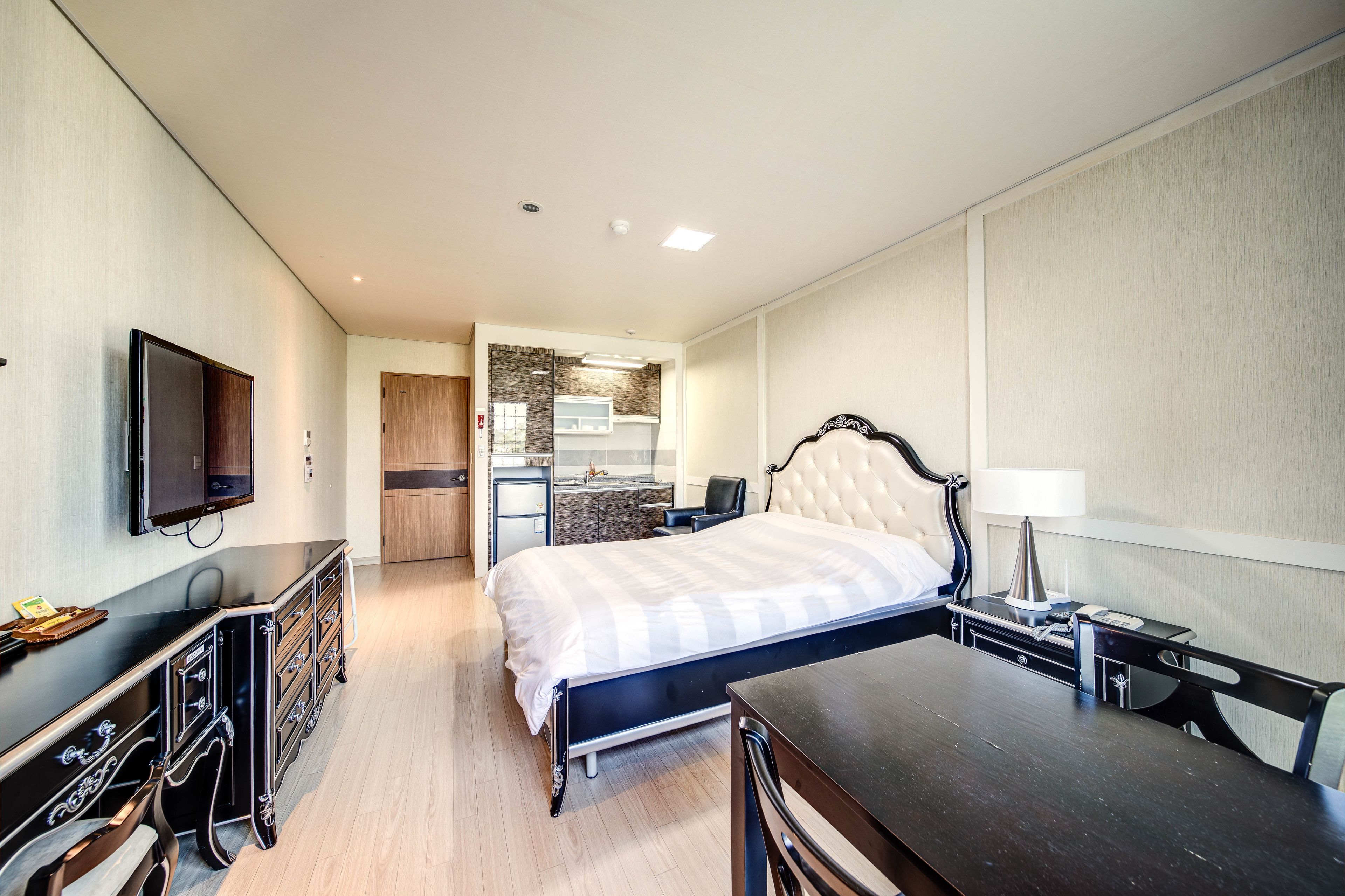 Habitación de hotel con cama, escritorio, silla, televisión y una kitchenette al fondo.