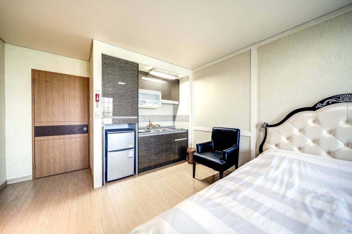 A hotel room with a bed, a chair, a kitchenette, and a wardrobe.