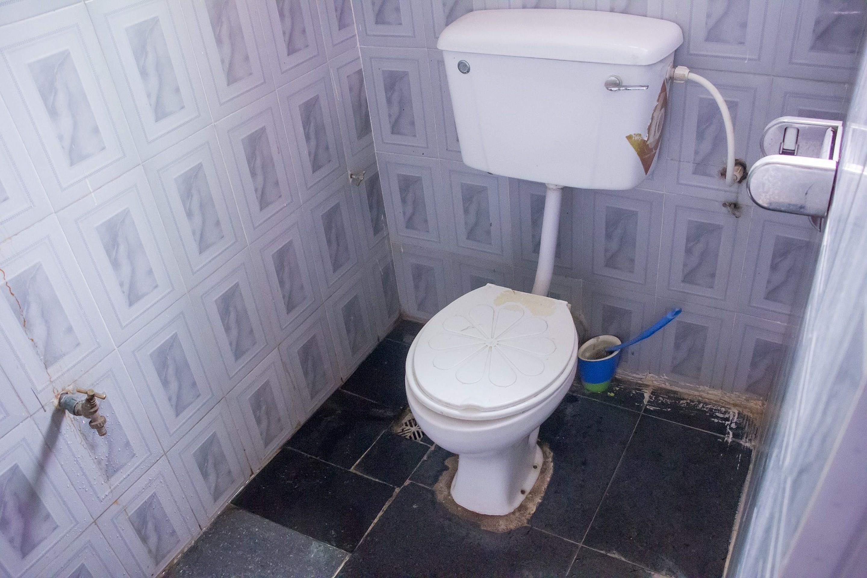Egy kis, egyszerű fürdőszoba, amelyben egy WC, egy kék vödör és egy kézi szivattyú található.