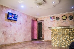 A hotel lobby with a TV mounted on the wall displaying a news channel, a reception desk with clocks showing different time zones, and a marble wall with a pink accent. - Bavidi Hotel (Lagos)