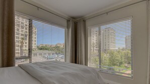 Habitación de hotel con una cama, dos ventanas con persianas, una cortina y vistas a edificios y árboles.