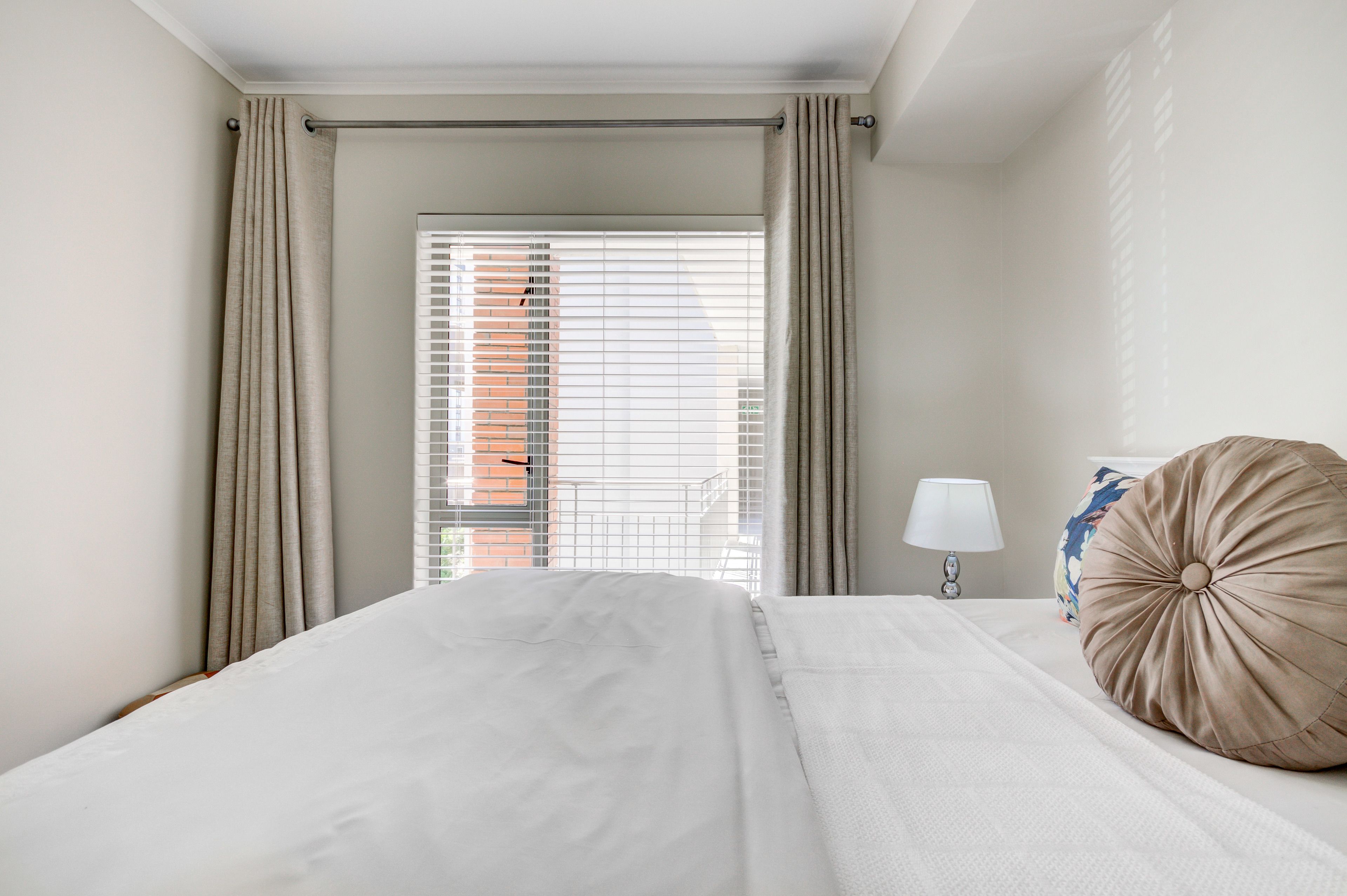 A hotel room with a bed, a window with blinds, a lamp, and a pillow.