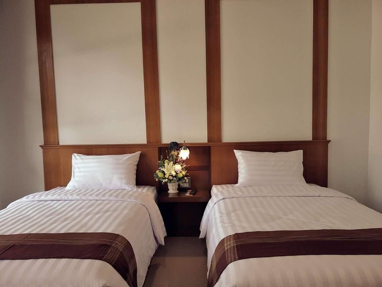 A hotel room with two single beds, wooden headboards, a nightstand with a lamp and flowers, and a plain wall.