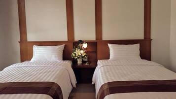 A hotel room with two single beds, wooden headboards, a nightstand with a lamp and flowers, and a plain wall.