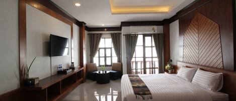 A modern hotel room with a large bed, a flat-screen TV, a seating area with a small table, and large windows with curtains.