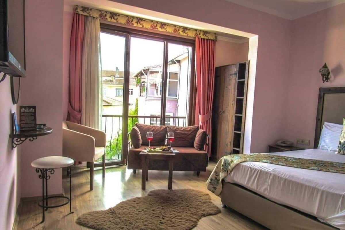 A bedroom with a bed, a sofa, a small table with wine glasses, a rug, and a window with curtains.