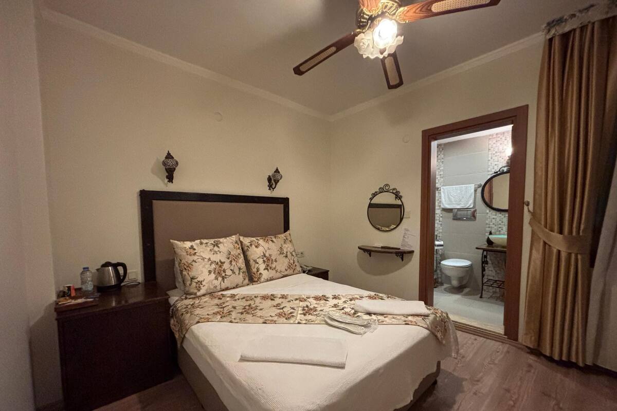 A neatly made bed with floral bedding, a bedside table with a kettle, a ceiling fan, and a bathroom visible through an open door.