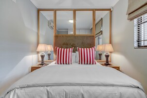 Una cama bien tendida con ropa blanca y almohadas a rayas rojas y blancas, flanqueada por dos lámparas de noche sobre mesitas de madera.