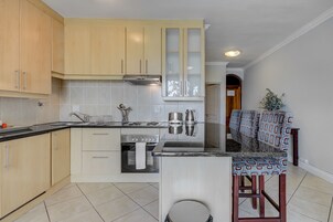 Una cocina moderna con armarios de madera clara, una estufa de acero inoxidable y un horno empotrado. Hay un taburete con un cojín estampado y una pequeña planta en maceta.
