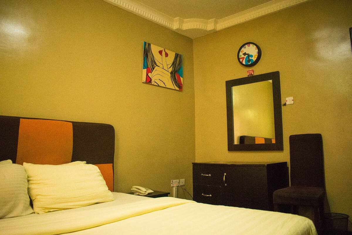 Een hotelkamer met een bed, een dressoir, een stoel, een schilderij en een klok aan de muur.