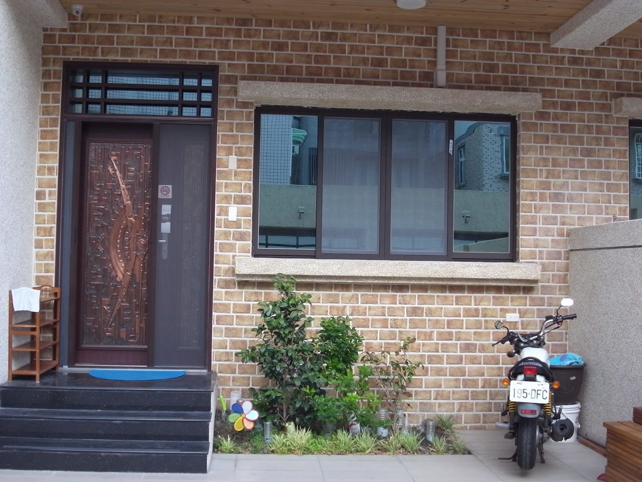 A brick house with a wooden door, a motorcycle parked outside, and a small garden with plants.
