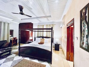 A hotel room with a four-poster bed, a ceiling fan, a desk with a chair, and a wall-mounted air conditioner. - Coconut Pointe Beach Resort (Prampram)