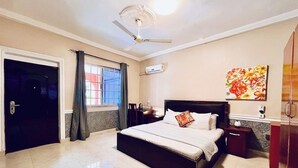 Une chambre à coucher comprenant un lit, un ventilateur de plafond, une fenêtre avec des rideaux, un climatiseur fixé au mur et un tableau.