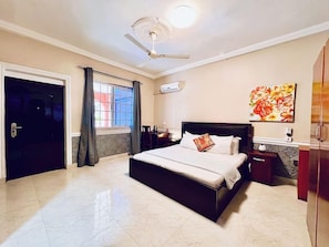 A bedroom with a bed, a ceiling fan, a window with curtains, a wall-mounted air conditioner, and a painting. - Coconut Pointe Beach Resort (Prampram)