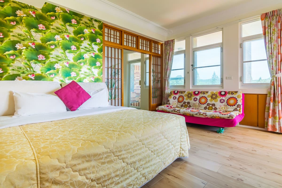 A bedroom with a bed, a colorful sofa, a decorative wall, and large windows.