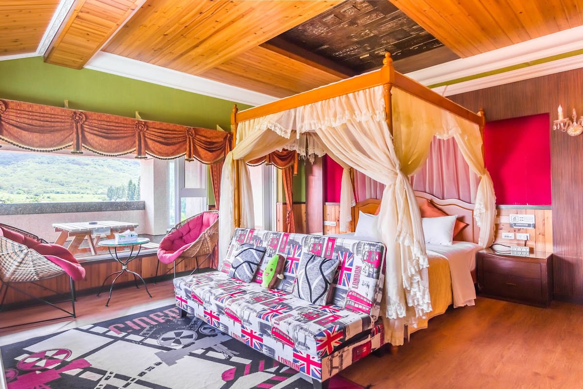 A bedroom with a four-poster canopy bed, a sofa with patterned cushions, a dining table, and a view of the outdoors.