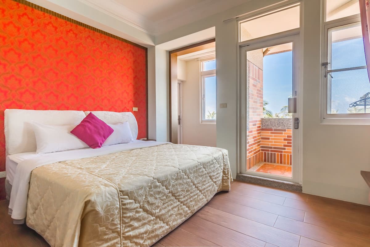 A hotel room with a bed, a red patterned wall, and a view of a brick building through an open door.