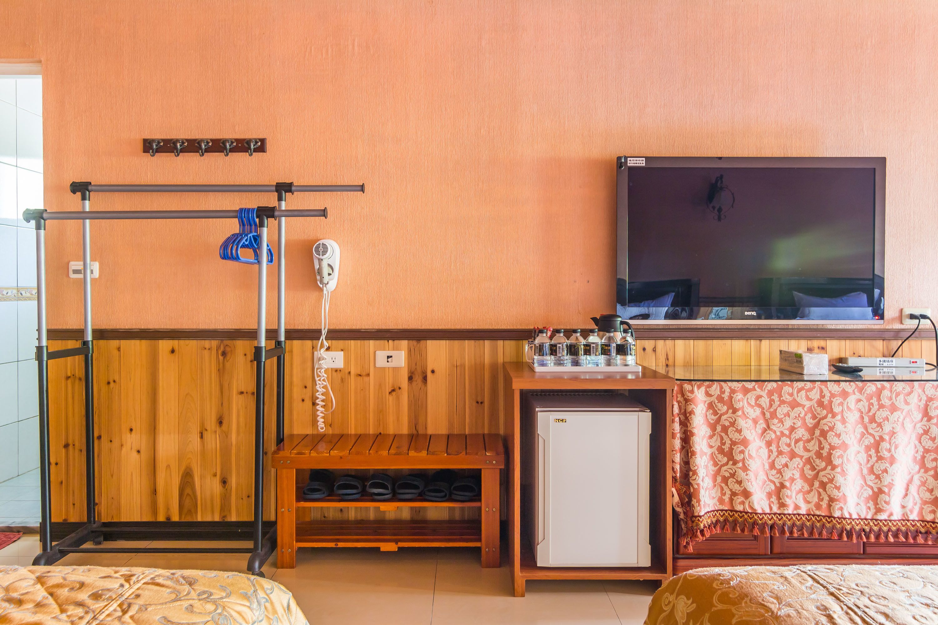 Habitación de hotel con un banco de madera, un refrigerador pequeño, un televisor montado en la pared y un zapatero.