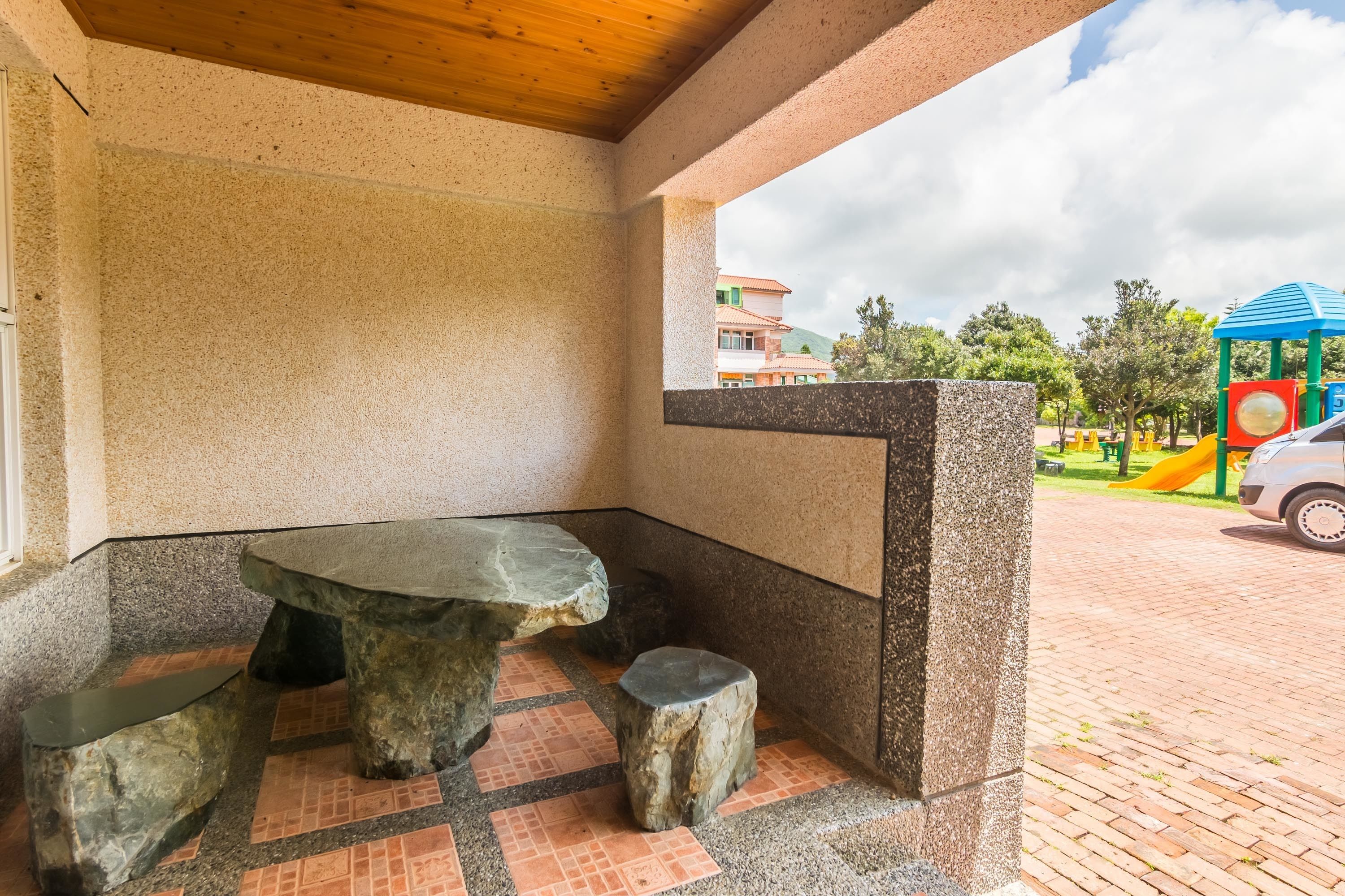 Un banco y una mesa de piedra en un espacio cubierto con vistas a un parque infantil.