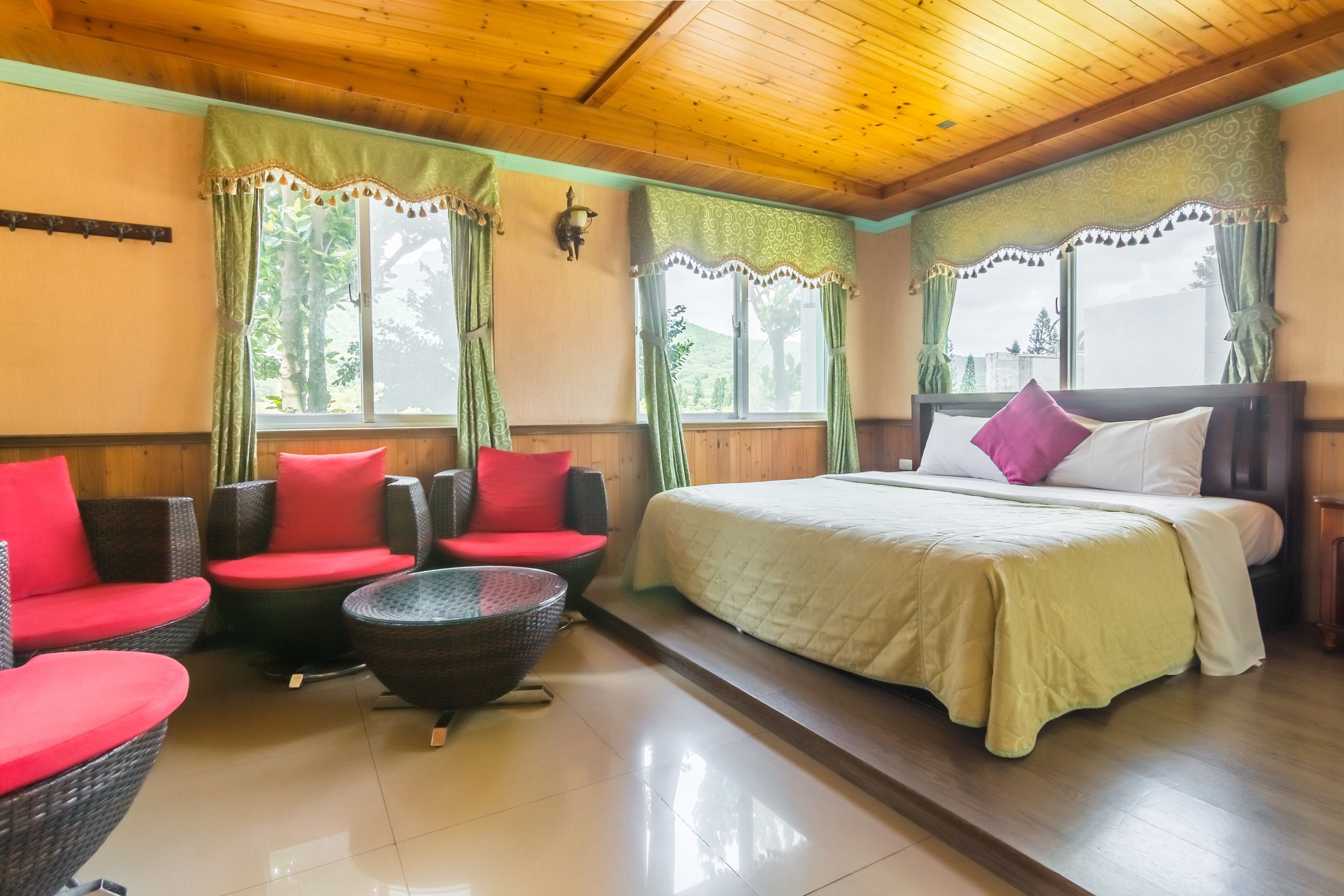 Una habitación con una cama, dos sillas rojas acolchadas, una mesita redonda y dos ventanas con cortinas verdes.