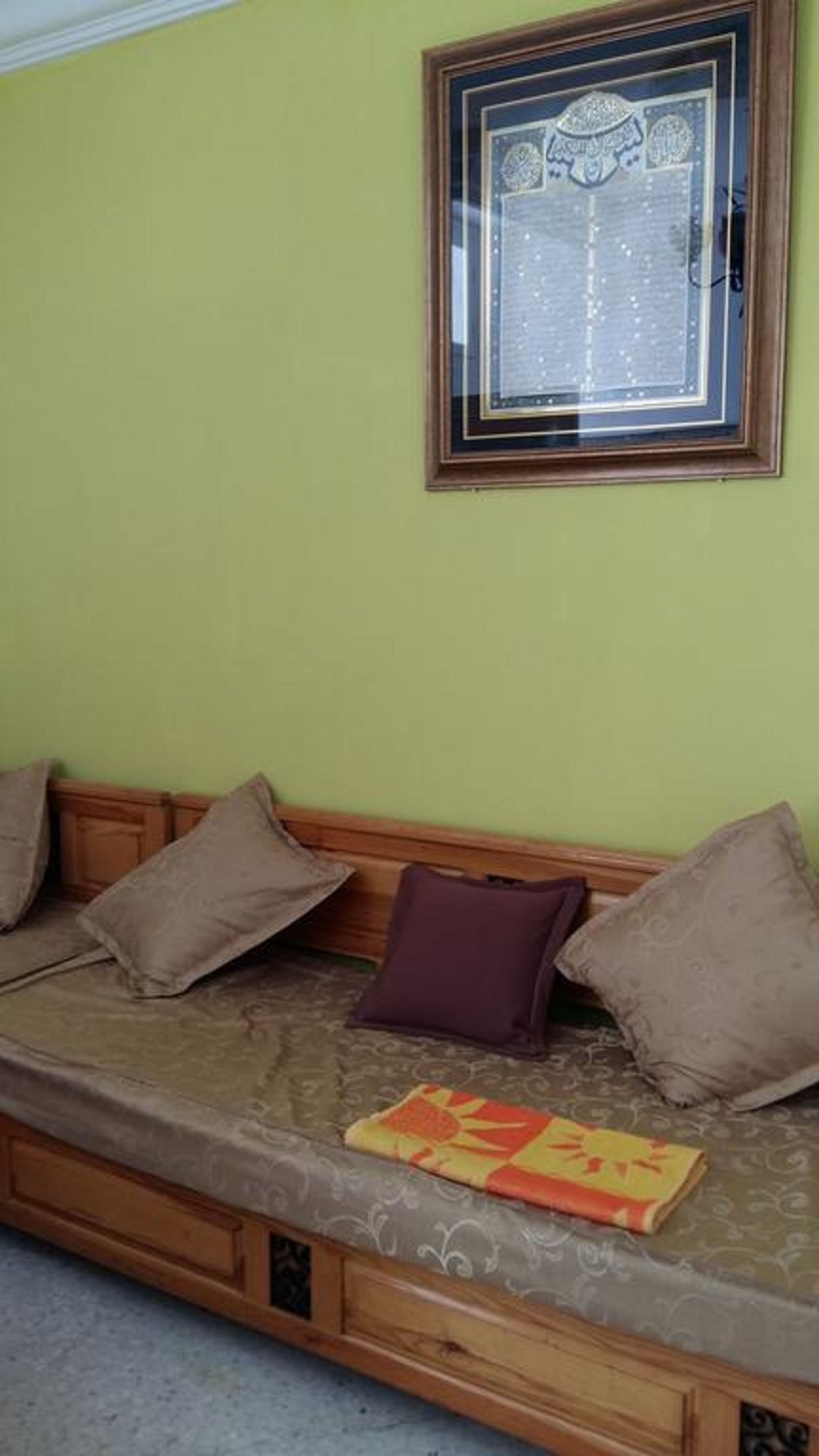 A wooden sofa with cushions against a green wall, a framed decorative piece above it, and a patterned cushion on the floor.