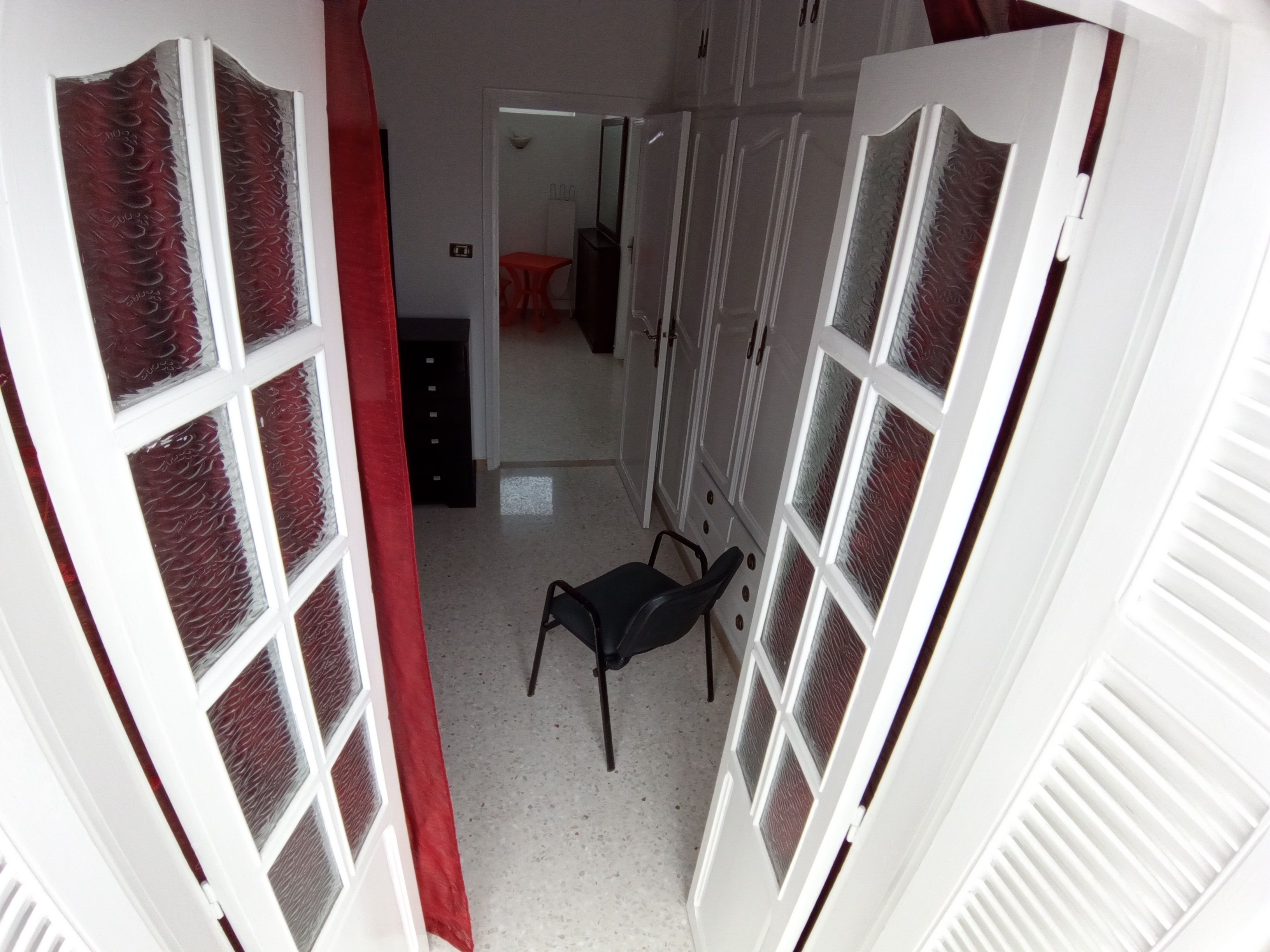 Un couloir étroit, avec des portes blanches et des rideaux rouges, qui mène à une pièce contenant une chaise et une petite table.