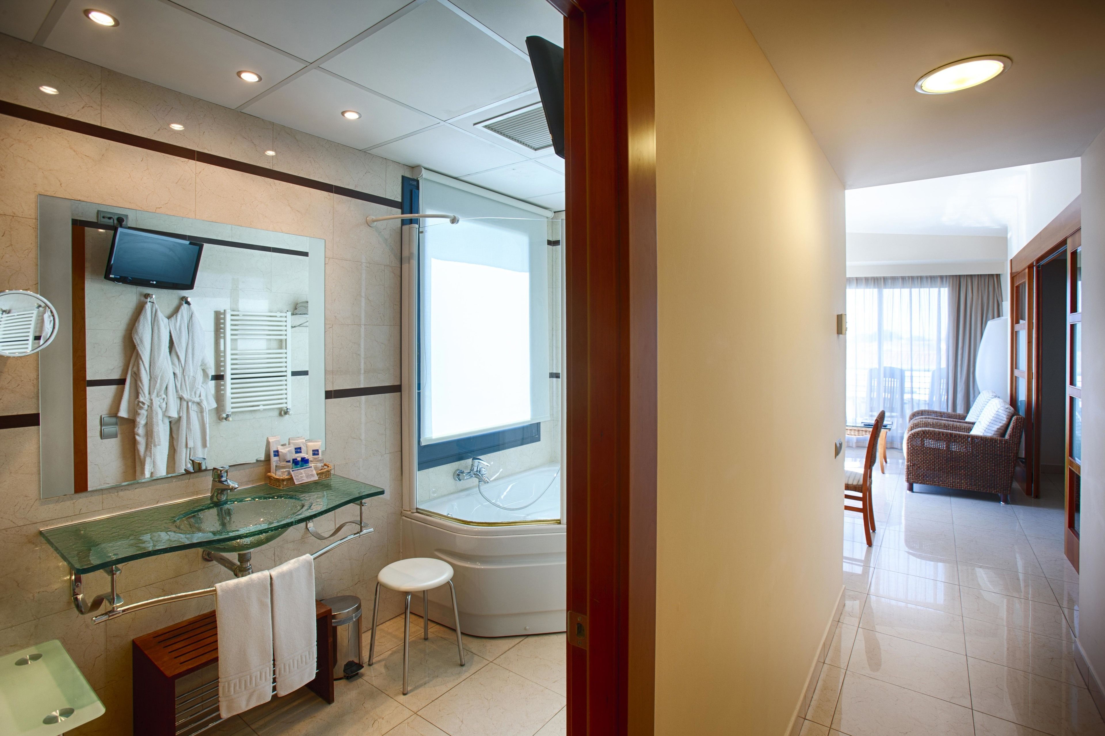 A modern bathroom with a glass sink, a bathtub, and a shower. There is a mirror with a TV, a towel rack, and a small stool. The bathroom leads to a living area with a sofa and a window.